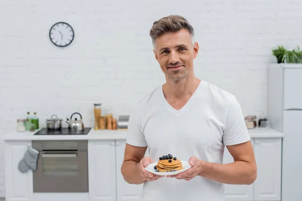 Uomo sorridente guardando la fotocamera e tenendo frittelle con mirtilli in cucina — Foto stock