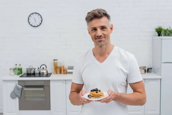 Sorrindo homem olhando para a câmera e segurando panquecas com mirtilos na cozinha — Fotografia de Stock