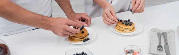 Ausgeschnittene Ansicht eines Mannes, der Blaubeeren auf Pfannkuchen in der Nähe von Frau und Honig in der Küche platziert, Banner — Stockfoto