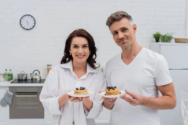 Smiling couple holding pancakes with blueberries and looking at camera at home — Stock Photo