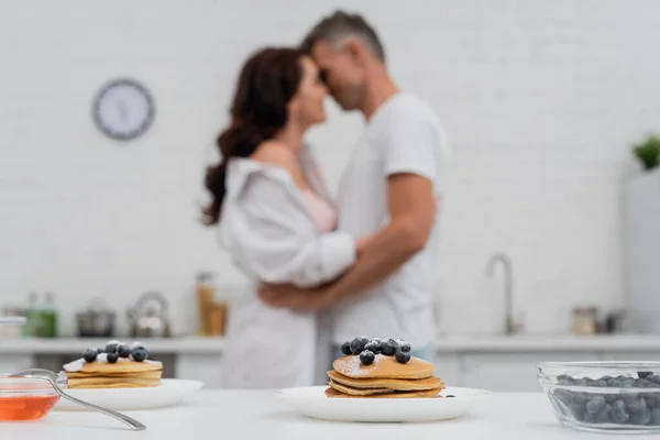Pancakes with blueberries near blurred couple kissing in kitchen — Stock Photo