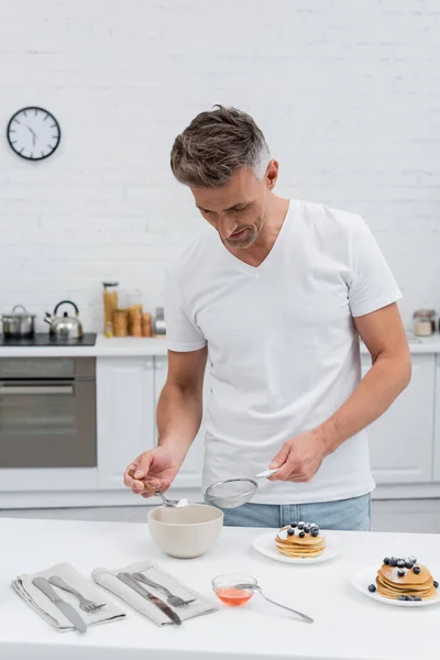 Hombre tomando azúcar en polvo de tazón y sosteniendo tamiz cerca de panqueques en la cocina - foto de stock