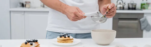 Vista cortada do homem segurando peneira com açúcar em pó perto de panquecas com bagas na cozinha, banner — Fotografia de Stock