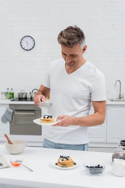 Smiling man pouring powdered sugar on delicious pancakes near coffee and honey in kitchen — Stock Photo