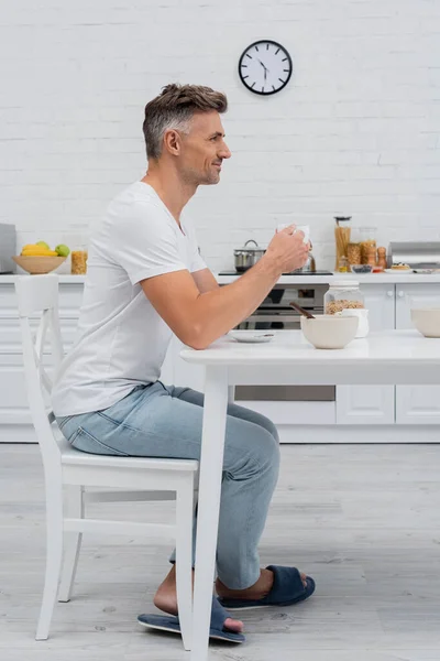 Vista lateral del hombre sonriente sosteniendo la taza de café cerca del desayuno en la cocina - foto de stock