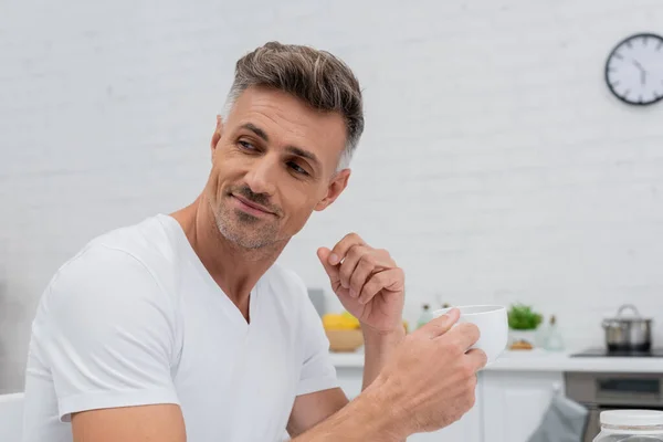 Hombre en camiseta sosteniendo taza de café en la cocina en casa - foto de stock