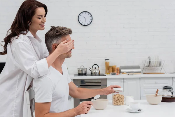 Femme souriante couvrant les yeux du mari près du petit déjeuner dans la cuisine — Photo de stock