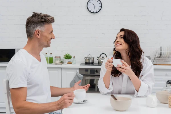 Smiling woman in shirt holding cup of coffee while husband talking near breakfast in kitchen — Stock Photo