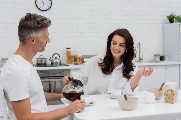 Mujer positiva hablando y vertiendo café cerca de marido y desayuno en la cocina - foto de stock