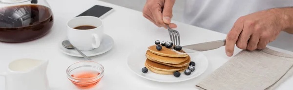 Cropped view of man cutting pancakes with blueberries near coffee and smartphone, banner — Stock Photo
