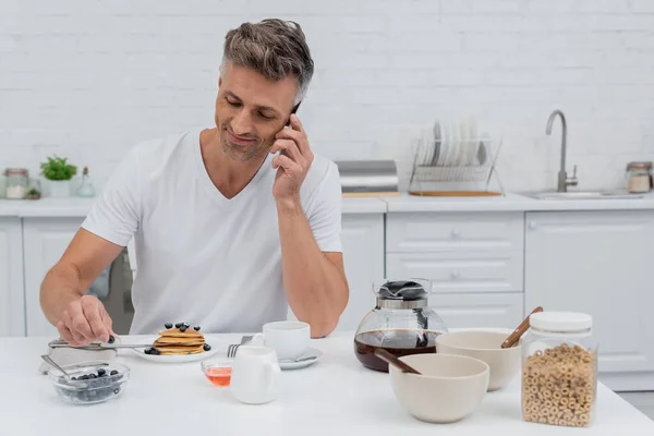 Sonriente hombre hablando en el teléfono inteligente cerca de delicioso desayuno en la cocina - foto de stock