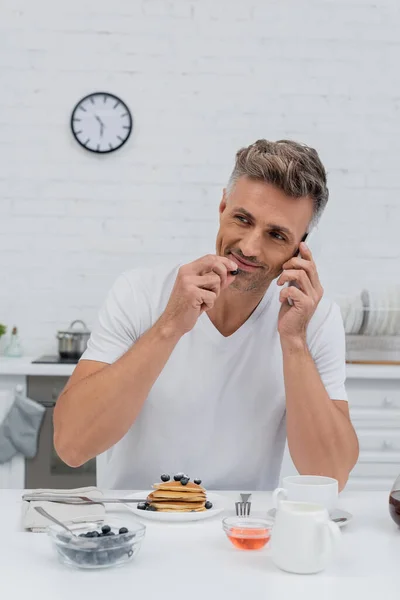 Man talking on cellphone and holding blueberry near pancakes and coffee in kitchen — Stock Photo