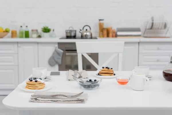 Вкусные блинчики с черникой рядом с кофе и смартфоном на кухне — стоковое фото