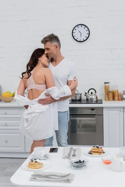 Vista lateral da mulher sexy em sutiã e camisa abraçando marido perto de café da manhã desfocado em casa — Fotografia de Stock