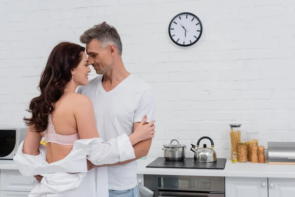 Vista lateral del hombre abrazando esposa sonriente en sujetador y camisa blanca en la cocina - foto de stock