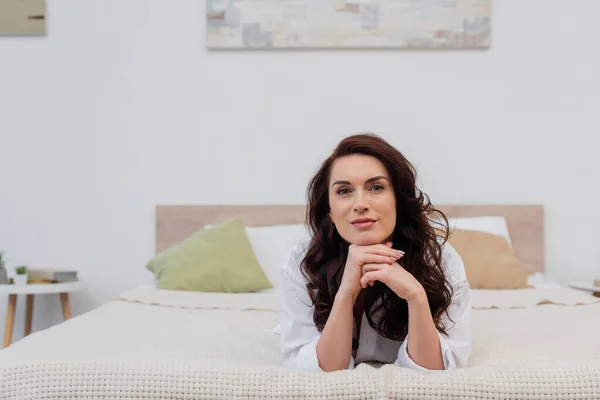 Brunette woman in shirt looking at camera while lying on bed — Stock Photo