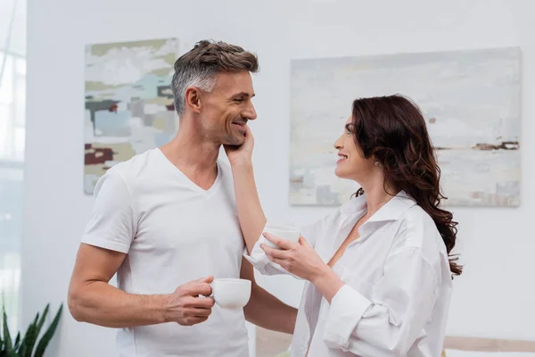 Vista lateral da mulher sorridente na camisa segurando xícara de café e tocando marido em casa — Stock Photo