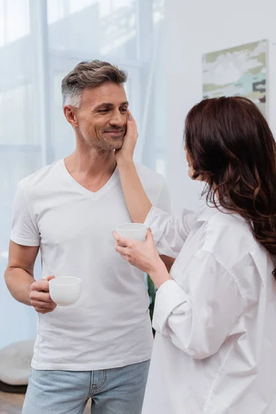 Brunette woman in shirt touching husband with cup of coffee at home — Stock Photo