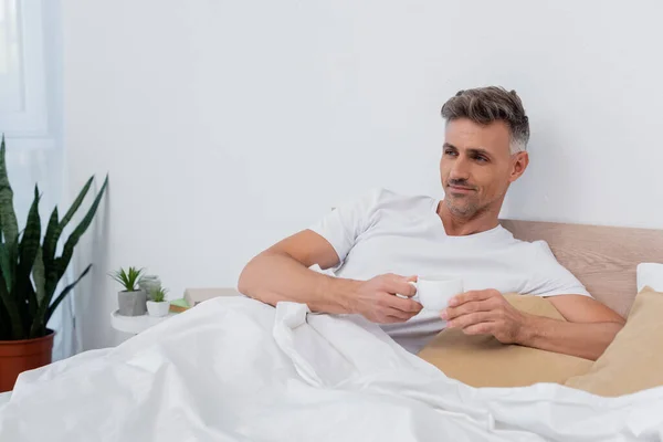 Mann im T-Shirt hält Tasse Kaffee, während er zu Hause im Bett liegt — Stockfoto