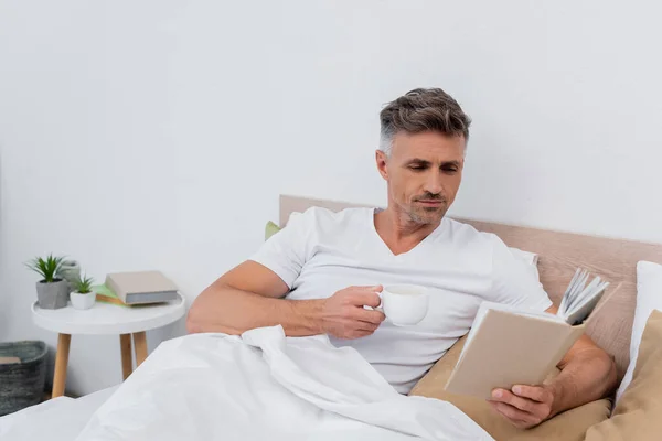 Man holding coffee cup and reading book on bed at home — Stock Photo
