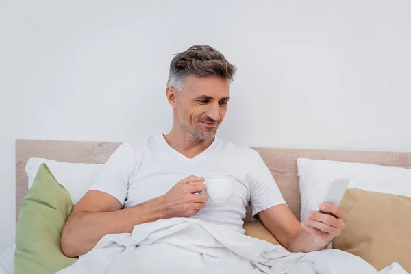 Hombre alegre usando teléfono inteligente y sosteniendo la taza de café en la cama en casa - foto de stock