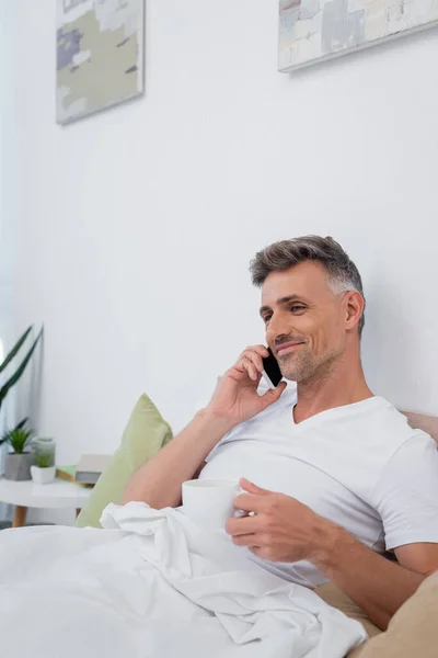 Sorrindo homem falando no celular e segurando xícara de café na cama em casa — Fotografia de Stock