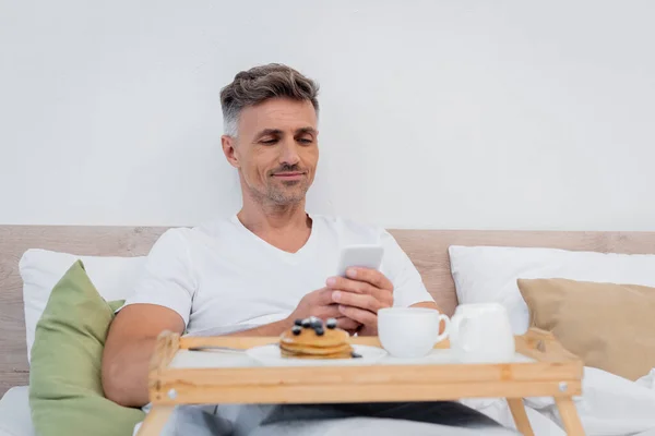 Мужчина пользуется смартфоном рядом с завтраком и кофе на кровати — стоковое фото