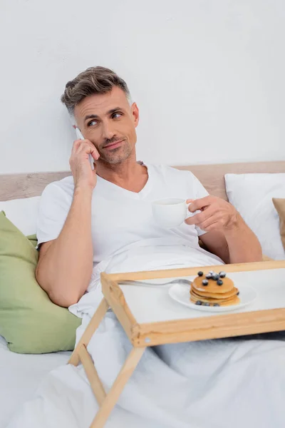 Man talking on smartphone and holding cup of coffee near blurred pancakes on tray on bed — Stock Photo