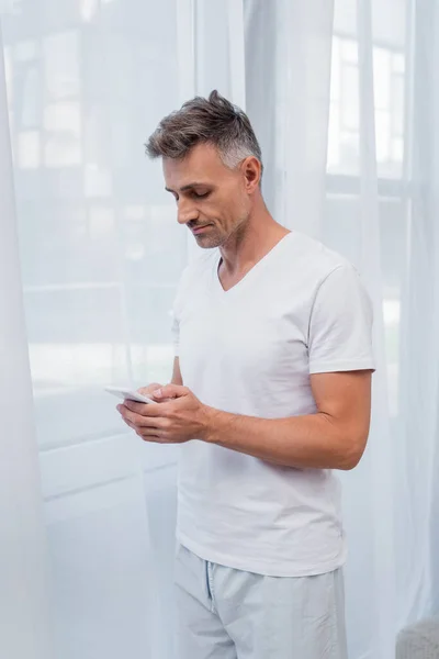 Homme en pyjama utilisant un téléphone portable près des rideaux à la maison — Photo de stock