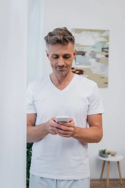 Улыбающийся мужчина в пижаме с помощью смартфона дома — стоковое фото