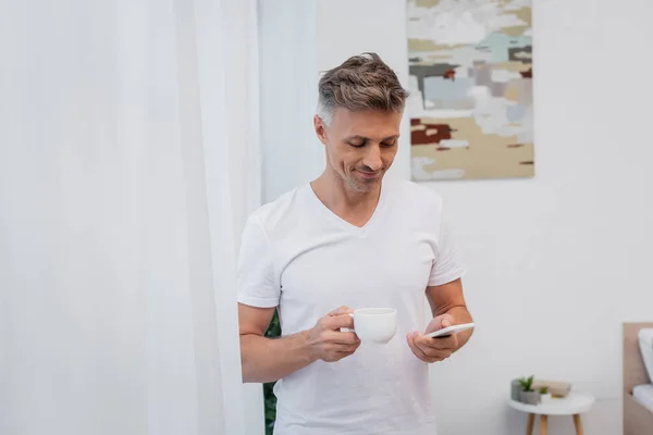 Homme positif en t-shirt tenant une tasse de café et utilisant un smartphone à la maison — Photo de stock