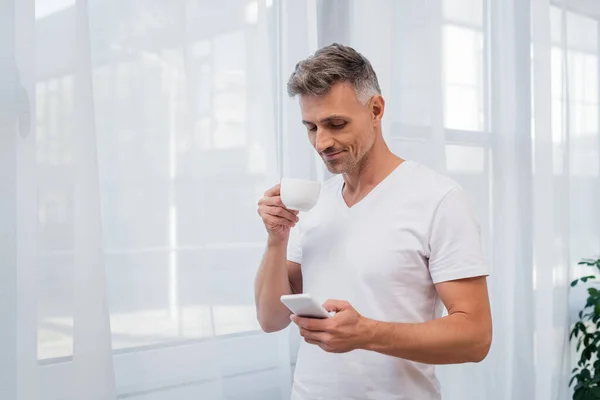 Uomo in t-shirt bianca con cellulare e tazza di caffè in mano a casa — Foto stock
