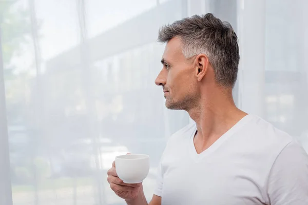 Vista laterale dell'uomo in t-shirt bianca con una tazza di caffè vicino alle tende a casa — Foto stock