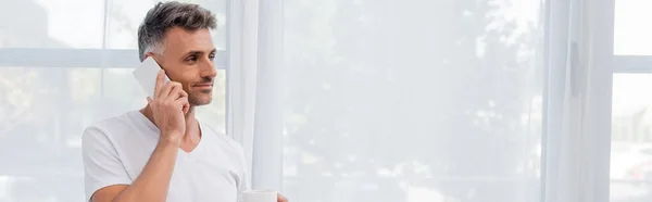 Hombre en camiseta blanca hablando en el teléfono inteligente y sosteniendo la taza en casa, pancarta - foto de stock