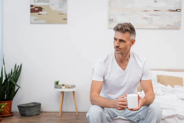 Мужчина в пижаме держит чашку кофе на кровати дома — стоковое фото