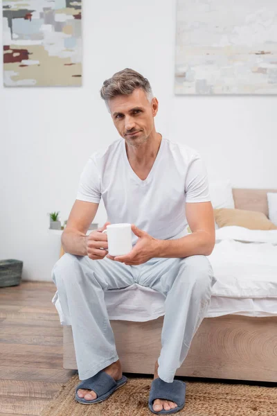 Man in pajamas and slippers holding cup of coffee while sitting on bed — Stock Photo