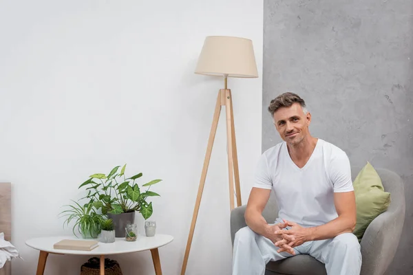 Homme souriant en pyjama regardant la caméra tout en étant assis sur un fauteuil à la maison — Photo de stock