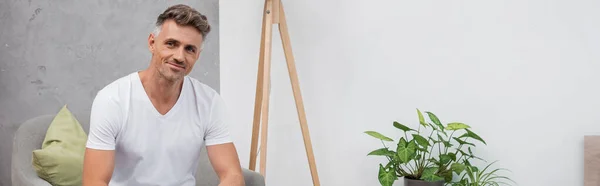 Positive man in pajamas looking at camera while sitting on armchair at home, banner — Stock Photo