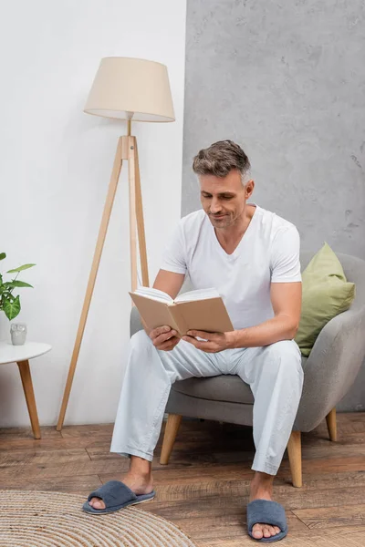 Man in pajamas and slippers reading book on armchair at home — Stock Photo