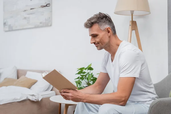 Vista laterale dell'uomo in pigiama lettura libro sulla poltrona in camera da letto — Foto stock