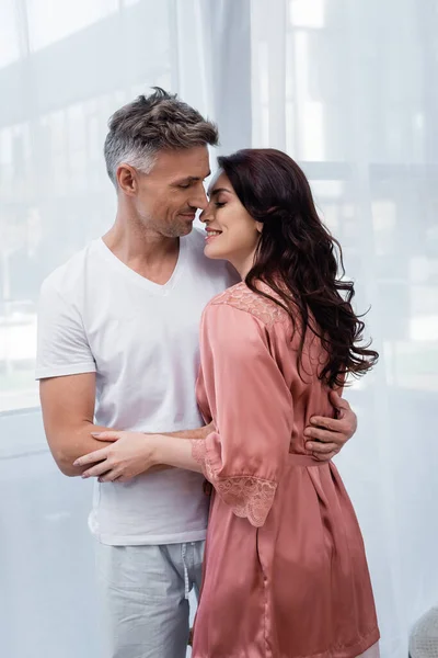 Homme en pyjama embrassant femme souriante en robe de soie à la maison — Photo de stock