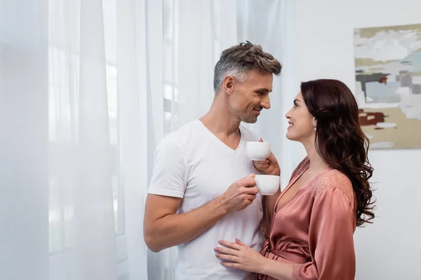 Vista lateral de casal sorrindo segurando xícaras de café em casa — Fotografia de Stock