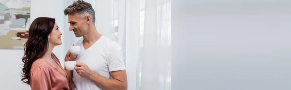 Vista lateral de mulher morena sorridente em robe de seda olhando para o marido com xícara de café em casa, banner — Fotografia de Stock