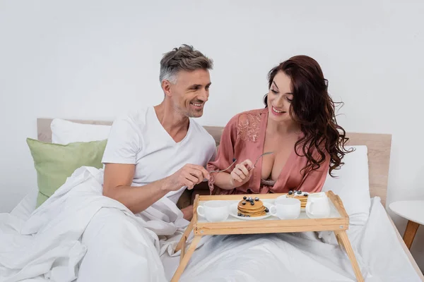 Coppia sorridente che tiene le forchette vicino a frittelle e caffè sul vassoio della colazione sul letto — Foto stock