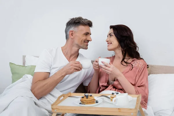 Smiling couple holding cups of coffee near blurred pancakes on tray in bedroom — Stock Photo