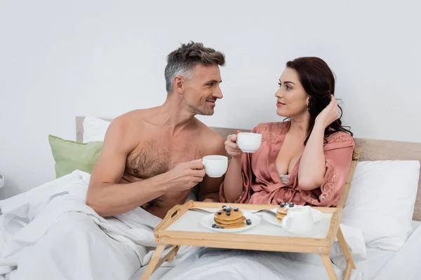 Hombre sin camisa sosteniendo taza de café cerca de la esposa en bata de seda y desayuno en bandeja en la cama - foto de stock