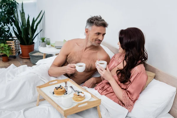 Улыбающийся мужчина без рубашки с чашкой кофе возле жены брюнетки и блинчиками на подносе в спальне — стоковое фото