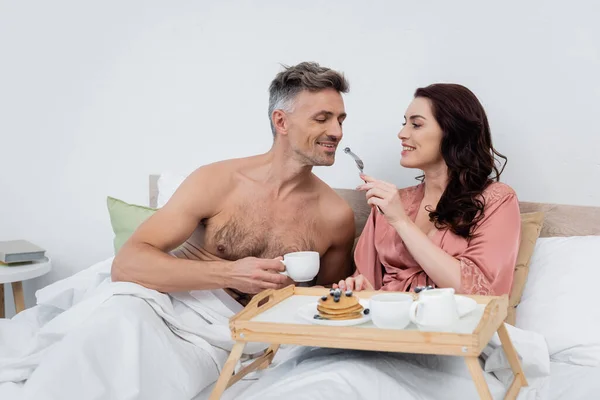 Cheerful woman in silk robe feeding husband with blueberry near breakfast on tray on bed — Stock Photo