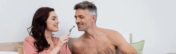 Smiling woman holding blueberry on fork while feeding shirtless husband on bed, banner — Stock Photo