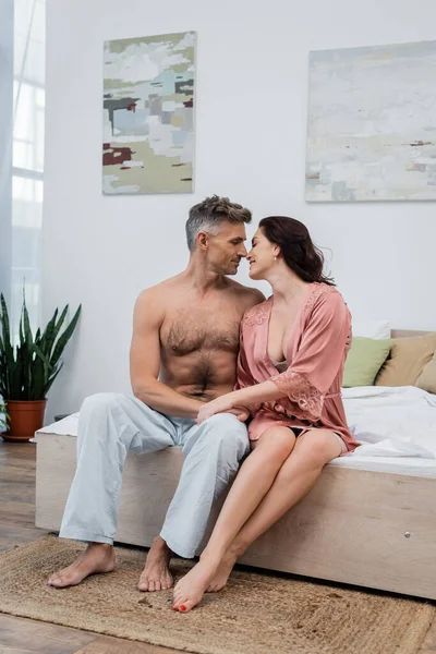 Side view of smiling woman in silk robe kissing shirtless husband on bed at home — Stock Photo